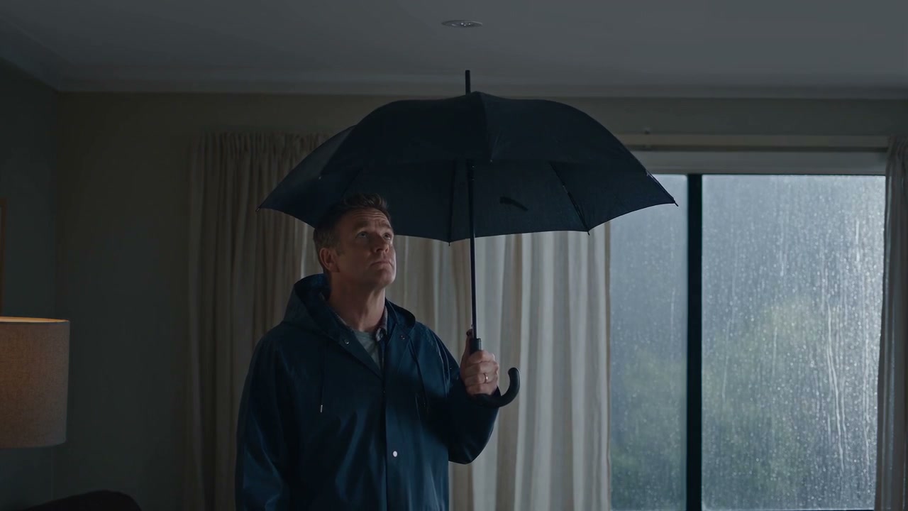 Rain streaming down a lounge room window while the owner stands inside holding an umbrella, from the film